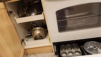 Open kitchen cabinet showing a selection of kitchen cookware including pots, pans, colander, and bakeware next to an oven.