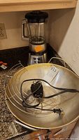 Electric wok metal bowl, wire stand, and power cord placed on kitchen counter next to Cuisinart blender base with glass pitcher lid