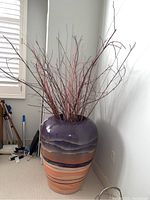 Full view of large decorative vase with dried branches placed on floor next to a window with plantation shutters.