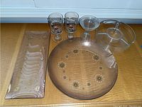 All items displayed on wood surface: two glass platters (one rectangular with floral and gold design, one round with flower motif), two stemmed glasses with flower decoration, glass serving platter with lid, and glass cup.