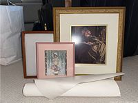 Photo showing two framed prints and one unframed rolled print on the floor. Larger gold framed print shows reclining woman; smaller pink framed print shows young girl with flowers. Rolled print is white paper.