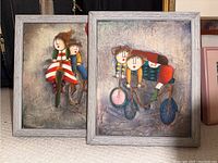 Two framed paintings shown together, children riding bicycles, signed, framed in gray wood frames.