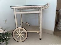 Side view showing large wicker wheels with spoke design and caster wheels, lower wicker shelf, and removable glass top.