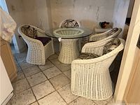 Four white wicker barrel chairs arranged around a glass-topped wicker table in a tiled room.