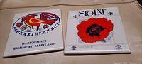 Photo showing two square ceramic hot plates on a fabric surface. Left plate features a colorful bird design with text 'Europa Imports Harborplace Baltimore, Maryland'. Right plate shows a red poppy flower with 'Stolat' text and decorative blue fleur-de-lis.
