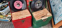 Two vintage metal storage boxes, one red and one green, each filled with 45 RPM vinyl records separated by tabbed folders. Each box has a handle and clasp closure.