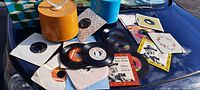 Wide view of collection of 45 RPM records on blue surface with orange record storage container and vintage record sleeves visible