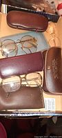 Top angled photo showing multiple vintage metal frame eyeglasses with original cases, including a brown case with printed advertising.