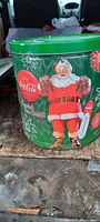 Side view of vintage Coca-Cola collector tin showing Santa Claus holding Coca-Cola bottles with green background and holiday snowflakes.