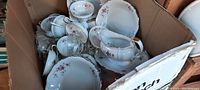 Full view of box containing multiple porcelain pieces including cups, plates, saucers, and gravy boat with floral pattern and gold trim.
