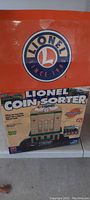Photo of the Lionel logo and boxed coin sorter showing front of the original packaging in orange and blue.