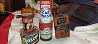 Photo showing 5 antique metal product cans: Calumet Baking Powder, Boscul Coffee, Gunk Car Wash, Esso Motor Oil, and Black Flag Insect Spray, arranged on a white cloth background with other items partially visible.