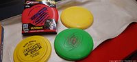 Four Frisbees arranged on a white surface, showing the sealed red and black Ultimate Frisbee, two yellow discs with different markings, and a green disc with a swirl design.