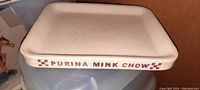 Side view of cream-colored rectangular stoneware feeder plate with red 'PURINA MINK CHOW' text and checkered accents on the rim.
