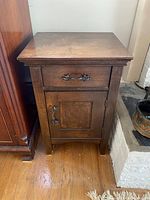 Front view of antique wooden end table showing a single drawer with metal handle and a cabinet door with matching handle, standing on a hardwood floor.