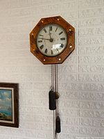 Full front view of octagonal wooden wall clock mounted on brick wall, showing weights and pendulum