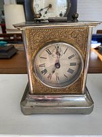 Front view showing the metal clock with detailed floral/scroll embossed design on the face surrounding the round dial with Roman numerals and a small seconds subdial.