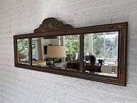 Full view of the antique wall mirror mounted on a textured white wall showing three mirror panels and ornate wooden frame with floral details and gold highlights.