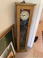 Full view of the tall antique Pulsynetic electric wall clock in oak case against a wall with some paintings behind.
