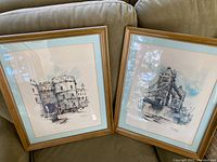 Two framed watercolors on a couch, showing detailed architectural scenes in wooden frames with blue matting.