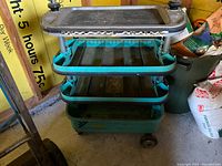 Full view of vintage metal tool caddy showing top tray with clamps and multiple lower teal trays for storage, mounted on wheels.