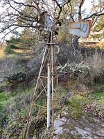 Full view of vintage wind turbine on rusty lattice stand in outdoor setting with trees and rocks.