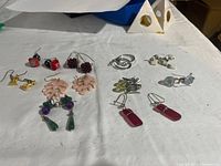 Ten pairs of assorted earrings laid out on a white surface, showing various shapes and colors including red cubes, purple clusters, coral pink florals, green drops, gold bird shapes, silver hoops, pearly whites, silver fish, and red rectangular drops.