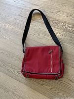 Photo of the front of the red messenger bag showing flap, zipper pocket and strap on wood floor.