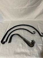 Five cast iron hooks varying in size and shape, laid out on white fabric. Three hooks with curling or twisted design shown (largest 16", medium 12", and smallest 9" pair).