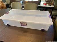 Clear plastic underbed storage container with purple latch and wheels shown closed on table.