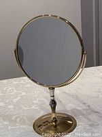 Front view of circular brass colored metal vanity mirror on a single stand with round base on a white patterned tablecloth.
