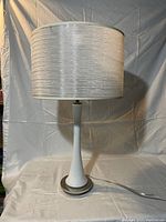 Full view of the lamp with shade, showing tall white ceramic base with silver metal accents and off-white textured fabric shade.