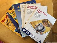 Seven packs of glossy photo printer paper arranged on a wooden surface showing various brands including two unopened Epson packs and other partially used packs.