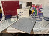 Photo showing an assortment of scrapbooking items including two red 12x12 albums, trimmer, cutting mat, precut paper shapes, page protectors, and fine tip pens.