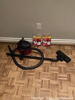 Photo of a red and black corded Shop Vac Micro Model MC150A including the main vacuum unit, attached hose, wand, floor nozzle, and two packages of vacuum bags placed on a parquet floor against a gray wall.