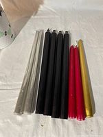 Ten taper candles in five silver glitter, four solid black, two red, and one gold laid on a white surface.