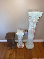 All three pillar pedestal stands, two light-colored classical style with rope tassels, one dark wood rectangular style.
