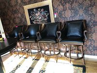 Set of 4 vinyl club style bar stools with black vinyl upholstery, carved wood twist-turned legs, nailhead trim, and cushions. Positioned in a row against a floral wallpapered wall near a door.