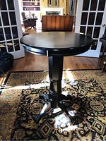 Black round bistro bar table placed on patterned carpet, with visible scratches and scuffs on the tabletop and base.