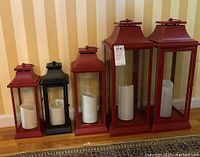 Five metal framed candle lanterns, four in red and one in black, each containing a white battery operated candle inside. Lanterns vary in height and are arranged in descending size order.