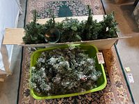 Top-down view showing green plastic storage bin filled with artificial Christmas garland with pinecones, and artificial tree branches on top.