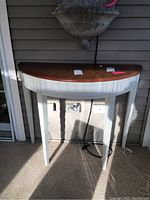 Frontal view of the half-moon shaped console table showing the brown stained top and white painted legs and base against a panel wall.