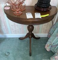 Frontal view of wooden demi lune side table showing semi-circular top holding glassware and decorative items, tri-foot pedestal base with carved details and claw feet.