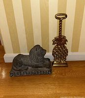 Lion door stop and brass pineapple door stop shown together on wooden floor against striped wallpaper.