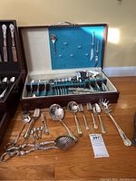 Photo showing open wooden case with various stainless steel utensils and flatware laid out, including spoons, forks, serving tongs, and large serving utensils with ornate handles.