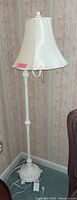 Full view of the white enameled metal floor lamp standing next to wallpapered wall.