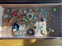 Full view of a wooden tray with assorted costume jewelry including bracelets, earrings, necklaces, and brooches arranged loosely.