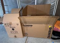Two cardboard boxes side by side on a concrete floor, one large labeled NuWave Bravo XL Smart Oven and one smaller labeled Keurig B70 WR