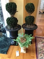 Front view of two large black urn-style planters with artificial topiary greenery and one smaller cream-colored planter with artificial foliage on a hardwood floor near a corner.