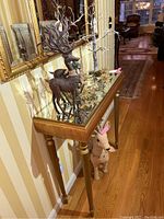 Shows two metal reindeer figures standing on a glass-top console table along with gold artificial foliage and a tall twig tree with lights. Below the table is a fabric reindeer figure.
